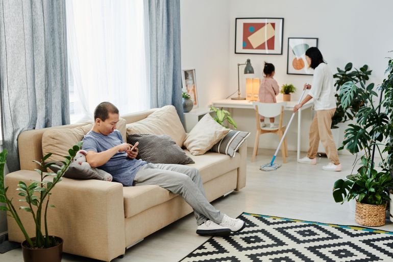 A family engages in daily chores, with a man relaxing and a woman mopping the floor.