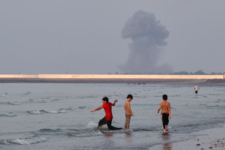 ap_6a56571eb3b0e-1784043294 Three boys play in the shallow waters of the Strait of Hormuz, as a plume of smoke rises from an explosion in the background, off Bandar Abbas, Iran, Monday, July 13, 2026. (Razieh Poudat/ISNA via AP)