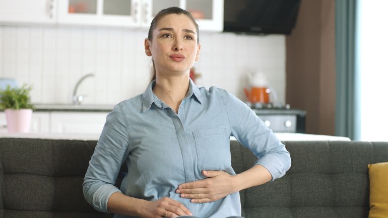 bloating2-1733405228 Young woman suffering from stomach spasm, abdominal pain at home. Stressed young woman massaging her belly on sofa, having stomach ache. Gastritis, abdominal or menstrual pain, diarrhea concept.