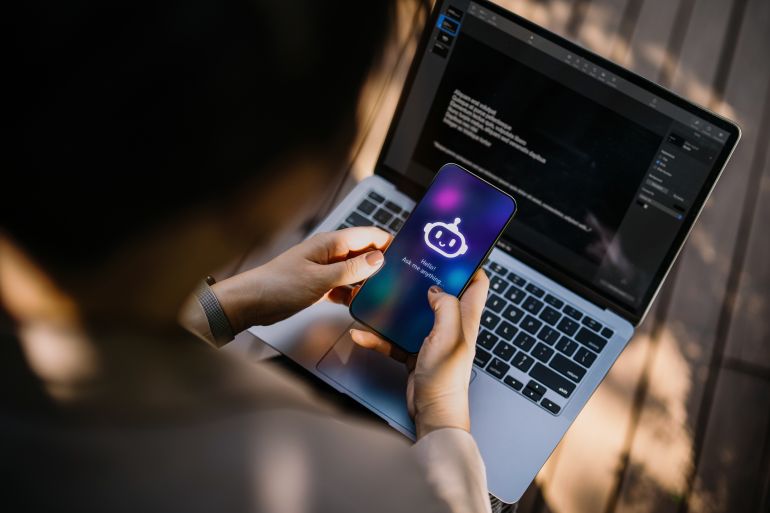 gettyimages-2193072141-1755598512 Businesswoman using chatting AI chatbot virtual assistance app on smartphone to assist her while working on laptop in office park. Using AI technology at work. Online robot system. Concept of VR and AR technologies. People, AI, innovation and technology