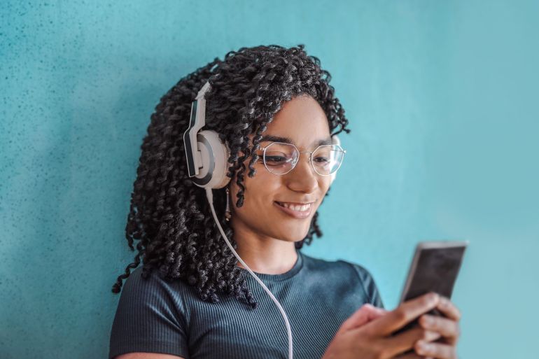 olly-3768125-1786457721 Portrait of a happy woman using headphones and smartphone against a blue wall.