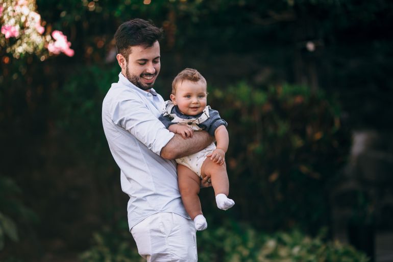 r-1785503115 Father joyfully holds his baby in a sunny park setting, capturing a moment of warm affection.