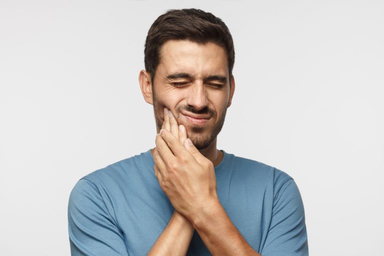 shutterstock_1190323033 Tooth ache. Young man feeling pain, holding his cheek with both hands, suffering from bad toothache, having painful expression