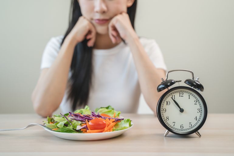 shutterstock_2491768389-1739704792 Intermittent fasting with clock, close up young woman, girl diet, waiting time to eat ketogenic low carb, green vegetable salad on plate. Eat food healthy first meal on brunch, lunch on table at home.