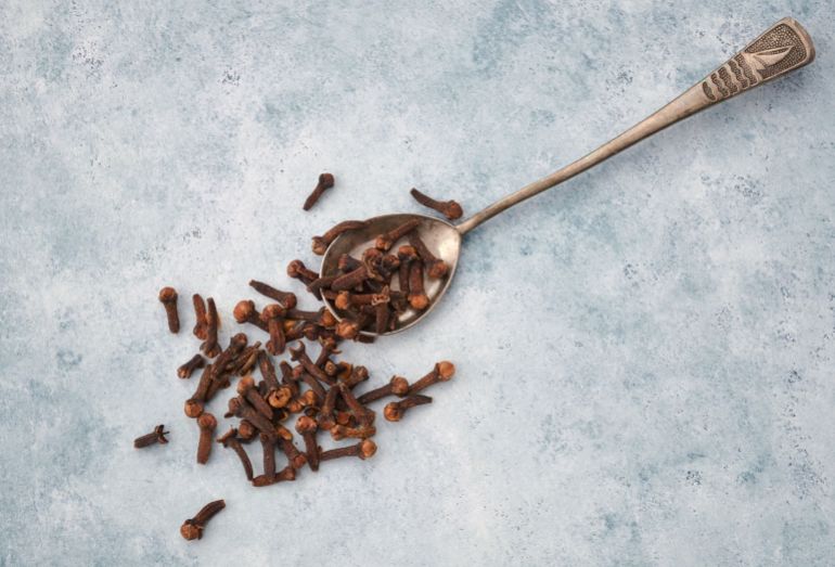049220-1788445469 Top view of aromatic cloves scattered beside a vintage silver spoon on a textured surface.