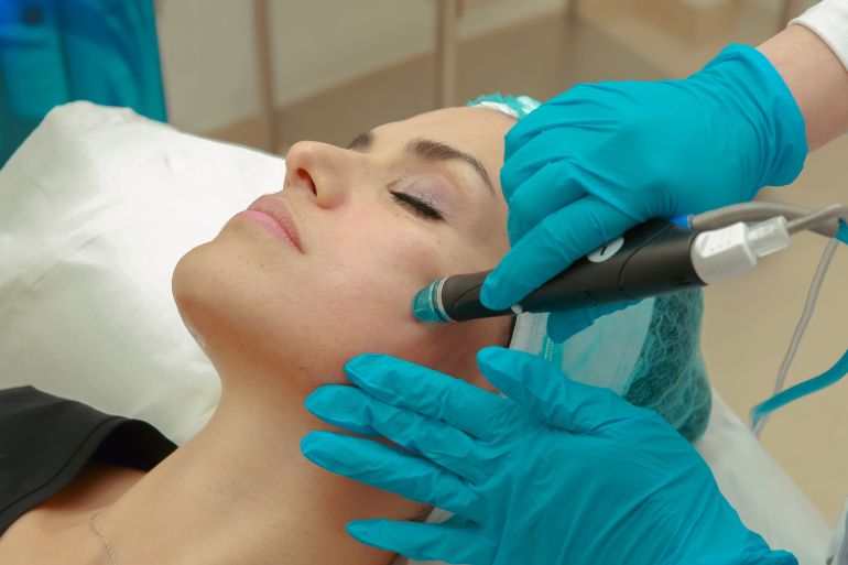 2556701-1788620479 A woman receiving a facial skincare treatment with a specialized device wearing protective gloves.