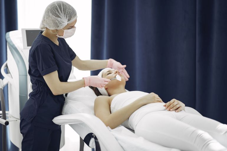 332-1787507342 Aesthetician applying facial treatment on a relaxed woman in a modern spa clinic setting.