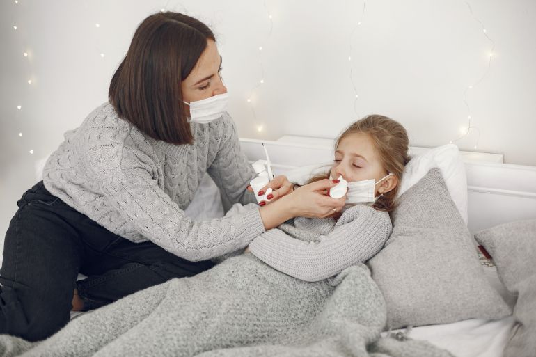 34479-1788696023 Mother administering nose drops to her sick daughter in a cozy bedroom setting.