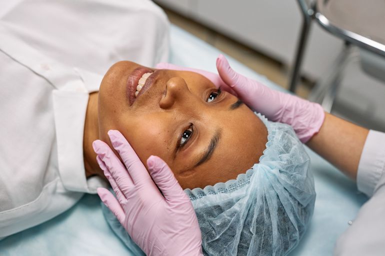 581081-1788625399 A woman receiving a facial treatment in a spa, wearing a hairnet while hands in gloves gently touch her face.