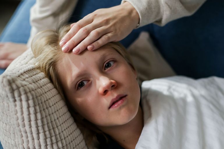 5858741-1788696533 Young child lying down with a hand checking for fever, indicating illness or flu symptoms.