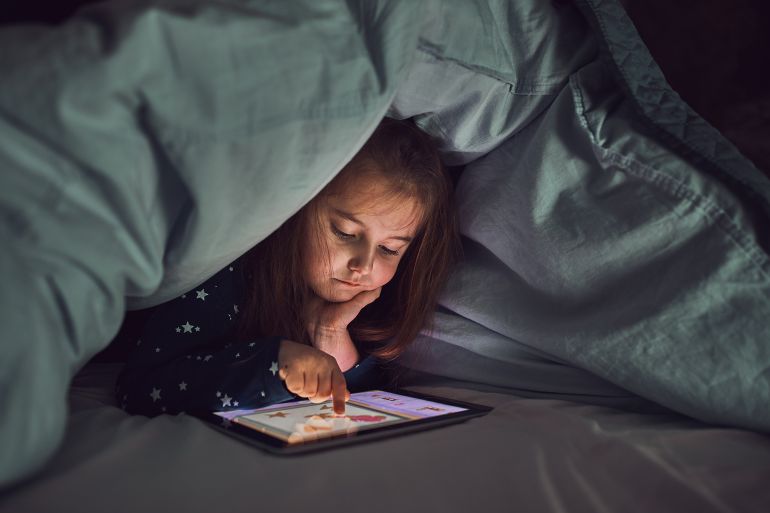 Little girl having fun watching, playing and listening to stories on tablet computer. Child having fun before bedtime laying under duvet using tablet by Przemek Klos @ادوبي ستوك