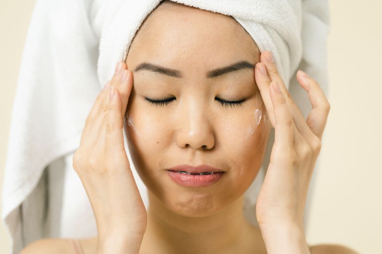 665-1787507140 Asian woman with a towel on head applying moisturizer for skincare routine.