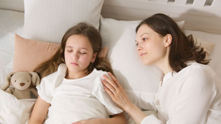 7504650-1788696783 A loving mother gently watches her daughter sleep peacefully in bed.