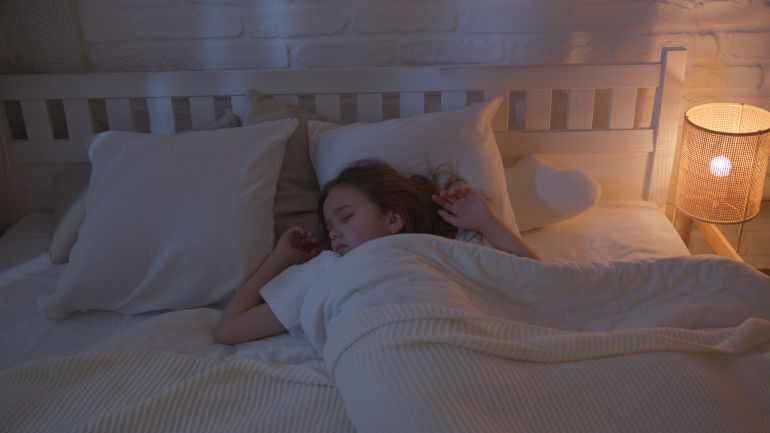 7504922-1788696917 A young girl peacefully sleeping in a cozy, dimly lit bedroom with a night lamp.