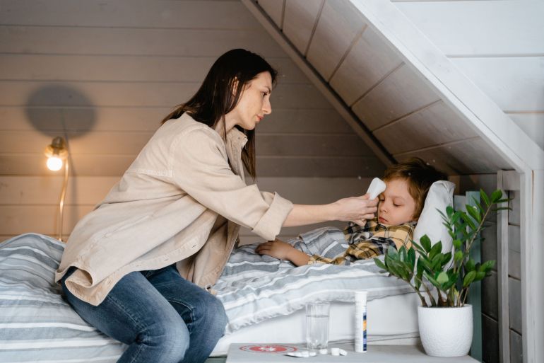 7641242-1788695933 A mother checks her sick child's temperature in a cozy bedroom setting, showcasing care and parenting.