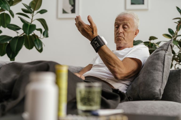 900026-1787583262 Elderly man checks his blood pressure using a wrist monitor in a comfortable living room setting.