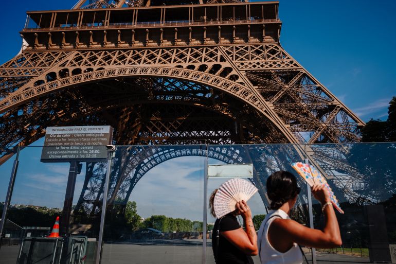 france-1787241278 Tourists protect from the sun under fans while walking next to the Eiffel Tower with a giant screen announcing its early closing due to heatwave in Paris on July 11, 2026. The operator of the Eiffel Tower said the latticed-steel Paris monument would "exceptionally close" early over the weekend at 4 pm (1400 GMT), as France was gripped by a third heatwave since May. (Photo by Dimitar DILKOFF / AFP)