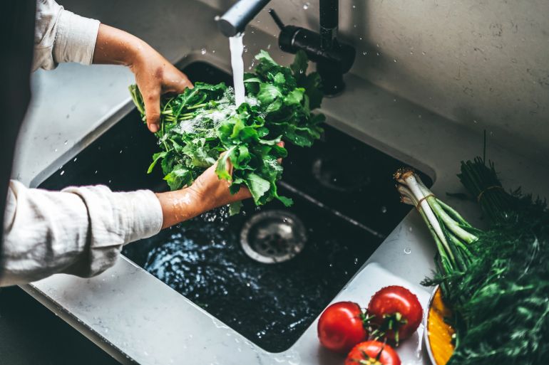Hands rinsing fresh green leafy vegetables under flowing water in the sink, ensuring cleanliness for healthy cooking. Bright kitchen scene with tomatoes and herbs adding vibrancy to the setting.