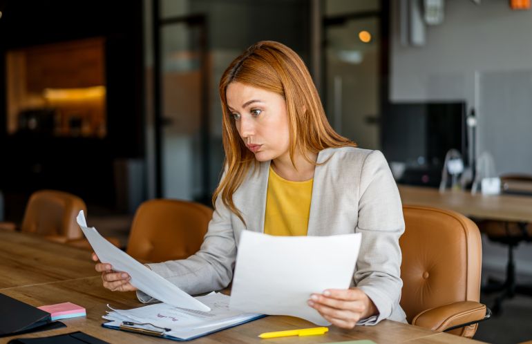 Young businesswoman working in modern office, carefully reviewing financial documents with a puzzled expression, analyzing company performance
