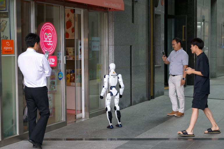 gettyimages-2285264097-1789197669 Pedestrians and onlookers gather to observe and film a Unitree R1 humanoid robot on a sidewalk in Singil, Yeongdeungpo-gu, Seoul, on June 24, 2026. The bipedal machine attracts public curiosity as it stands outside a storefront, illustrating the growing presence of high-tech robotics in urban areas. (Photo by Chris Jung/NurPhoto via Getty Images)