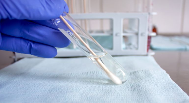 shutterstock_1699027549 "Cracow/Poland - April 2020: A hand with a protective glove holding a transparent glass test tube with cotton-tipped applicator with a swab sample in an extraction solution for medical test."