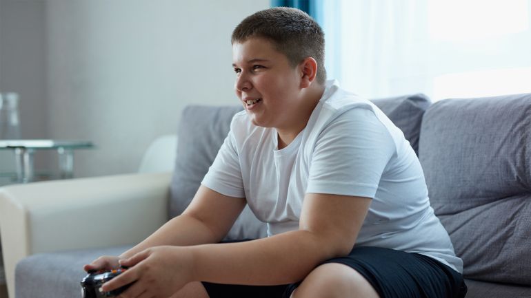 shutterstock_1801107010-1684875362 caucasian teenager boy enjoys video games, overweight boy sits on sofa holding console in hands, concentrated on game, look at screen shutterstock_1801107010
