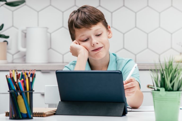shutterstock_2261895403-1776069656 Portrait of young preteen pretty boy wearing blue T-shirt, sitting at desk in front of digital tablet, writing drawing on screen with white pen stylus. Remote education, online school, e-learning.