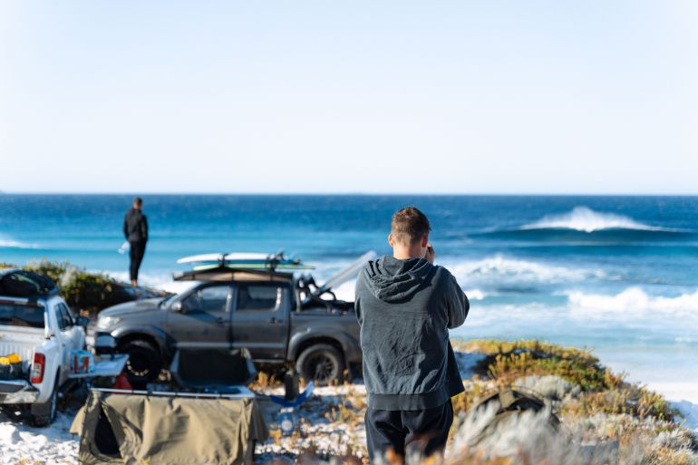 11351812-1791560424 Two men enjoying a beach day with trucks, surfboards, and ocean waves.