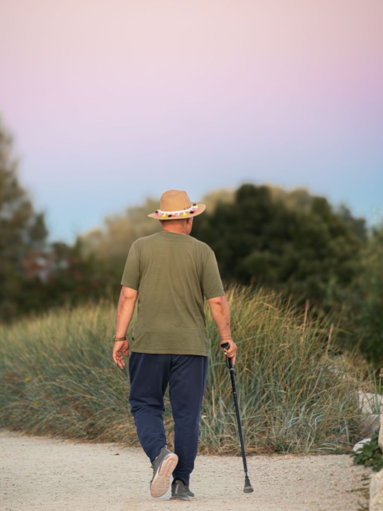 19808060-1790679534 Man in green shirt and hat walking on a rural path with a stick at sunset.