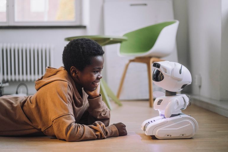 2ai-1791185098 Smiling boy talking with AI robot while lying on floor in innovation lab - stock photo
