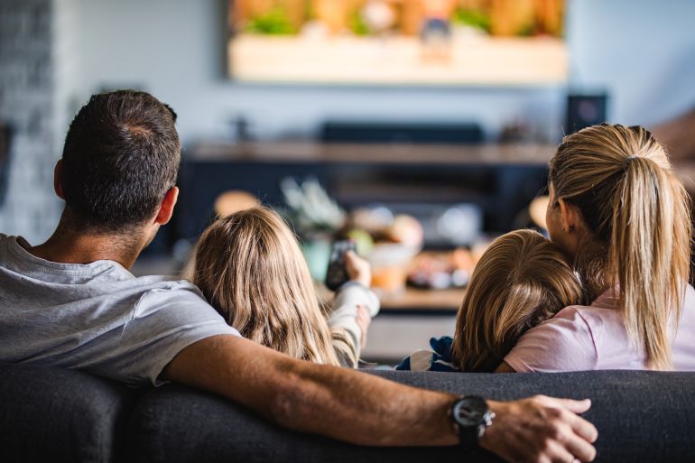 7654765-5-1793273504 Back view of a relaxed family watching TV on sofa in the living room.