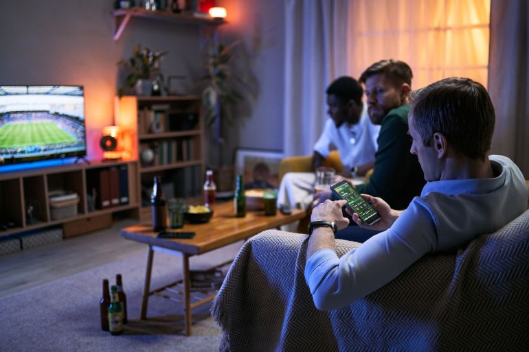 7654765-7-1793273519 Young adult male friends watching football match at home, one man checking smartphone betting app while socializing. Concept for male friendship, sports viewing, mobile wagering marketing