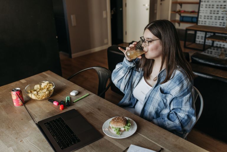 7801189-1790675299 A young woman drinks from a glass while working on her laptop with snacks nearby.
