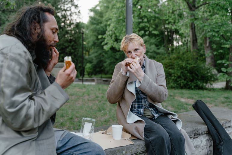 8078569-1790675572 Two individuals share a meal outdoors on a park bench surrounded by greenery.