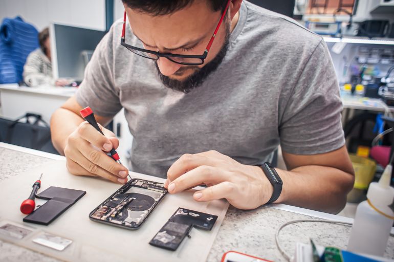 adobestock_555663154-1790590441 Workplace top view, close-up. Repairman disassembles smartphone with screwdriver in an electronics repair shop. المصدر AdobeStock