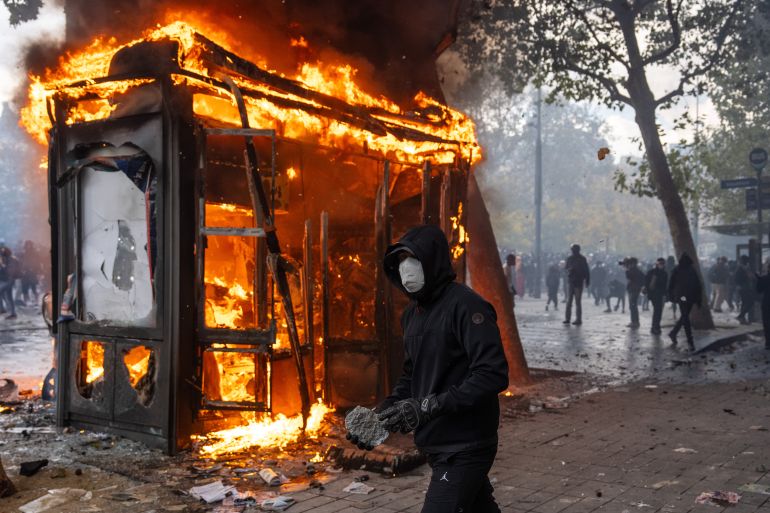 getty_6ad5eee6f3-1792405223 PARIS, FRANCE - OCTOBER 17: A protester holds a rock to throw at police as a kiosk burns during a demonstration near Place de la République, as the the Gilets Jaunes join students for a weekend of action on October 17, 2026 in Paris, France. The involvement of the Gilets Jaunes (Yellow Vests) with the ongoing French school blockades expands the scope of these demonstrations by linking localised student grievances to broader economic policies. Student unions originally initiated the movement to protest underfunding, deteriorating infrastructure, and acute teacher shortages across French high schools and universities. By joining the protests, the Gilets Jaunes introduce their established platform against austerity cuts, rising living costs, and surging fuel prices into the youth-led action. (Photo by Carl Court/Getty Images)