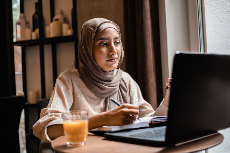 gettyimages-1451261997-1791915961 Portrait of cheerful muslim woman working online on laptop and making notes in notebook. Cafe space.