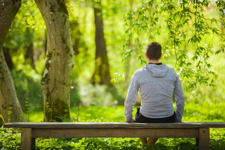 gettyimages-2152005382-1793046676 Young adult man sitting on bench and staring at green leaves, grass and trees. Thinking about life at beautiful park in sunny spring day. Spending time alone in nature. Peaceful atmosphere. Back view.
