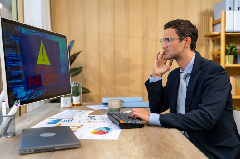Adult businessman is looking concerned at a computer monitor displaying a system hacked warning, showing his company's vulnerability to data breaches and cybersecurity threats