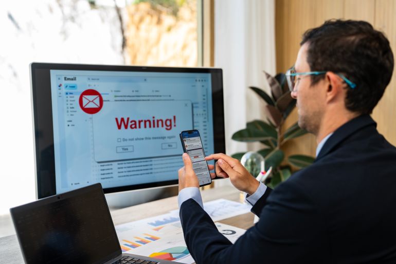 Businessman checking smartphone messages while facing a computer monitor displaying a warning! pop-up for an suspicious email, indicating a potential cybersecurity threat or phishing scam