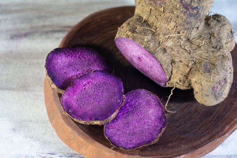gettyimages-2291232969-1792692024 A close up shot of a cut tuber root and raw purple yam ube slices showing bright purple flesh on a wooden plate