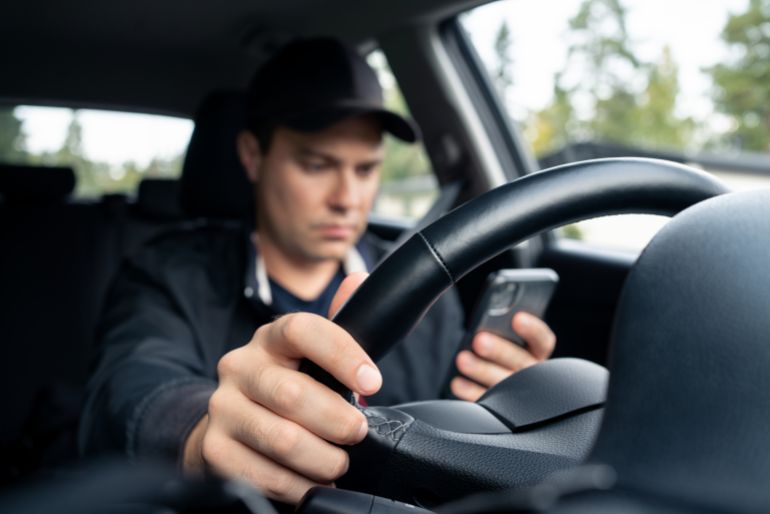 shutterstock_2282407525-1754222051 Driving car and using phone to text. Driver using cellphone. Accident, crash and danger in traffic. Man texting with mobile app. Distracted by mobilephone. Checking sms message with smartphone.